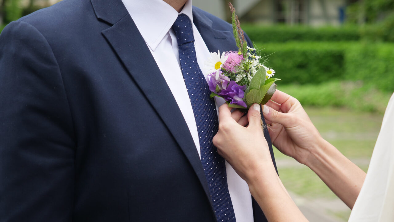 Nahaufnahme von zwei Händen, die ein kleines Blumensträusschen am Anzug eines Mannes befestigen. Der Mann trägt ein weisses Hemd und eine dunkelblaue Krawatte mit feinen, weissen Pünktchen.