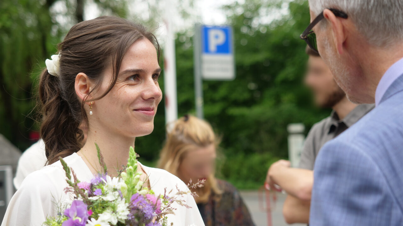 Leicht seitliche Aufnahme einer Frau mit Blumenstrauss in weissem Kleid, die lächelnd nach rechts zu einem Mann schaut, dessen Gesicht nicht zu erkennen ist. Im Hintergrund sind unscharf grüne Büsche, zwei Hochzeitsgäste sowie eine offene Barrierenschranke und ein Parkplatzschild zu erkennen. 