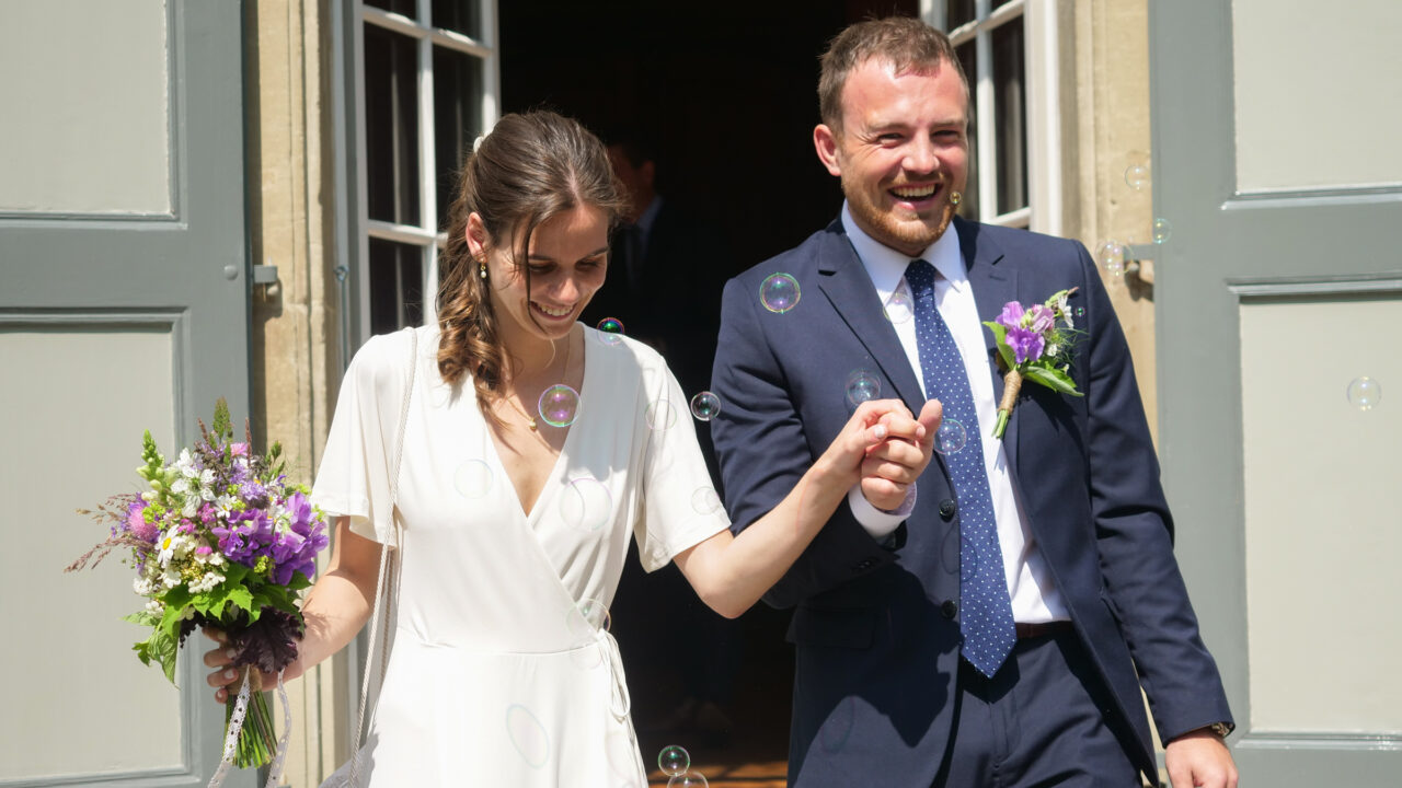 Frontalaufnahme einer Frau in weissem Kleid mit Blumenstrauss und eines Mannes in dunkelblauem Anzug, die Hand in Hand eine Treppe heruntergehen, die im Bild nicht zu sehen ist. Ein paar Seifenblasen tanzen um die zwei herum.