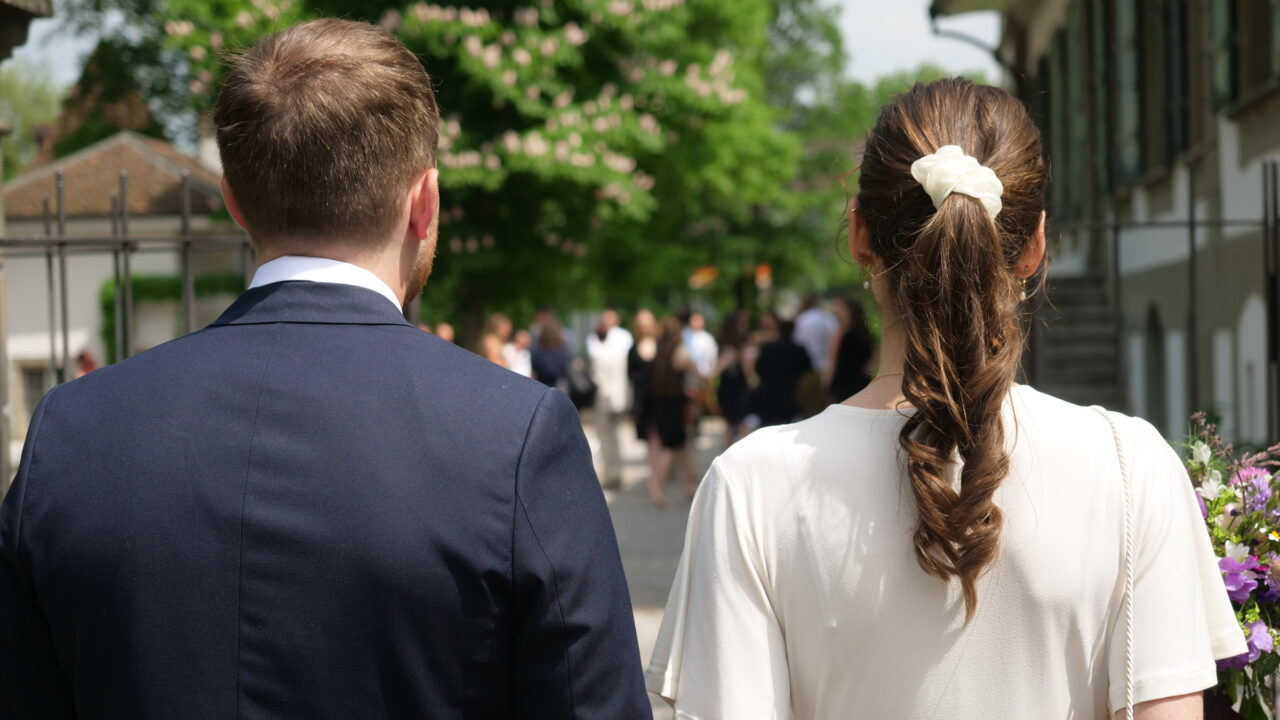 Aufnahme des Brautpaars von hinten, der Mann trägt einen dunkelblauen Anzug, die Frau ein weisses Kleid und mit einem weissen Gummi zusammengebundene Haare. Im Hintergrund ist unscharf eine Hochzeitsgesellschaft auf dem Kiesplatz vor dem Schloss Bümpliz.