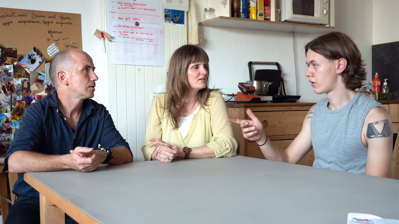 Zwei Männer und eine Frau sitzen in einem Büro an einem Tisch und diskutieren