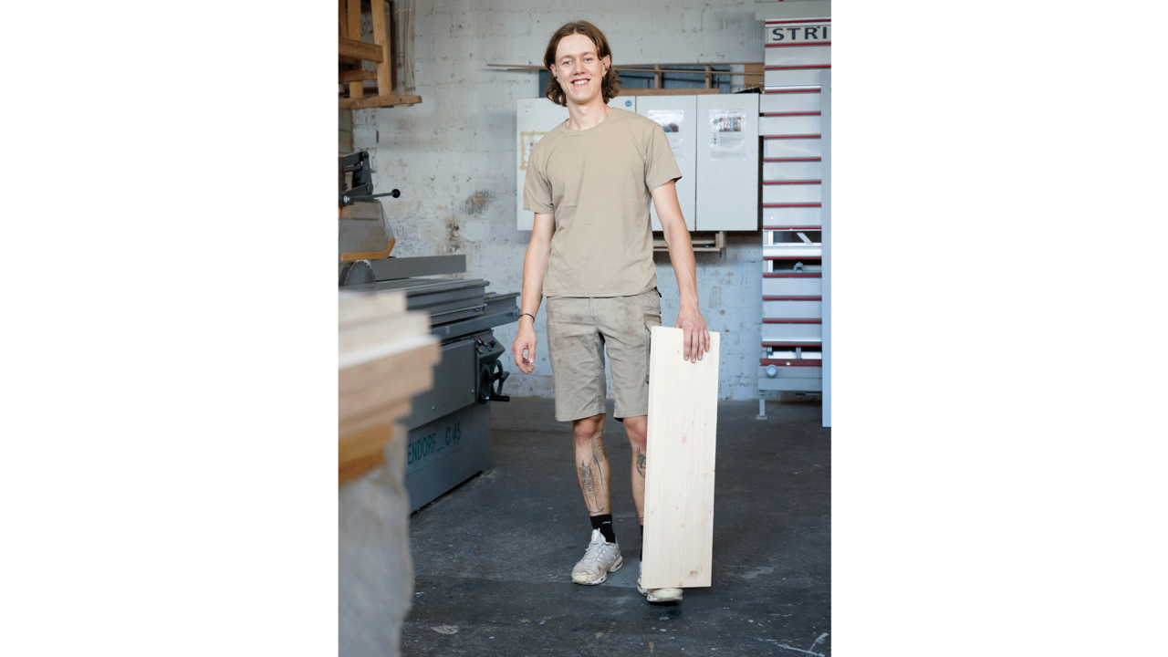 Ein junger Mann steht frontal zur Kamera, mit der Hand hält er ein Holzbrett fest, das auf seinen Fuss abgestützt ist