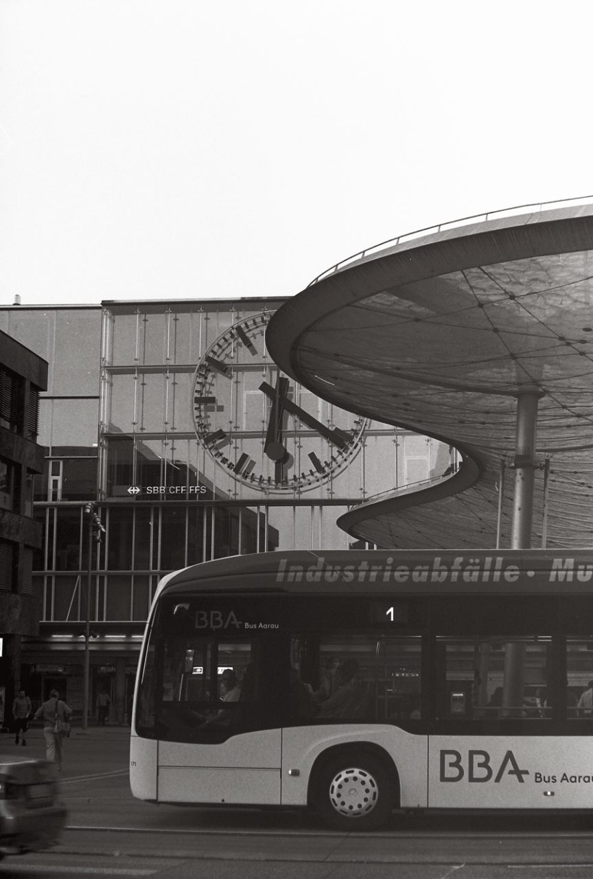 Schwarzweissfotografie vom Busbahnhof Aarau. Auf dem Bild sieht man das Wolkendach, die grosse Bahnhofsuhr und einen Bus.