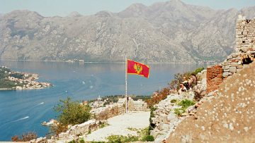 Aussicht über die Bucht von Kotor in Montenegro mit einer rot-goldenen Nationalflagge im Vordergrund, umgeben von Bergen, Wasser und historischen Steinruinen.